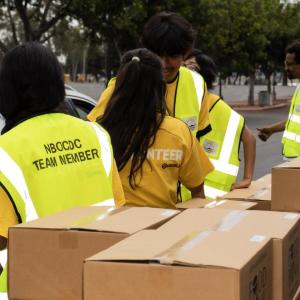 Volunteers in yellow vests gather around a long table stacked with sealed brown food bank boxes.
