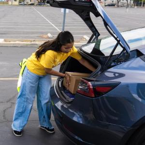 A volunteer in a yellow shirt places a brown box in the trunk of a blue car in a parking lot.