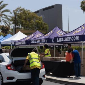 A food distribution site with LA Galaxy-branded canopies and volunteers loading boxes into a white sedan.