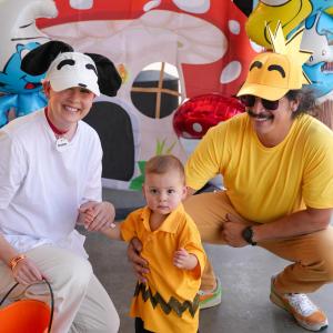 Two adults in character costumes pose with a young child dressed in an orange-and-black Halloween costume beside colorful decorations and an orange trick-or-treat bucket.