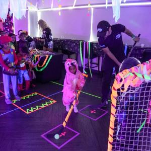 A child in a pink costume uses a hockey stick during a neon-lit indoor hockey activity as other costumed children wait nearby.