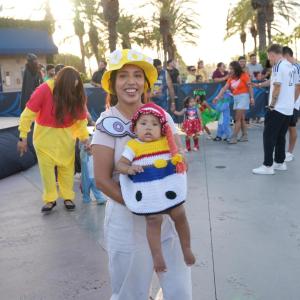 An adult holds a baby dressed in a colorful Halloween costume as families in costumes gather outdoors beneath palm trees.