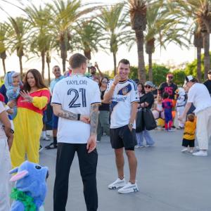 LA Galaxy players mingle with families in Halloween costumes during an outdoor community celebration lined with palm trees.