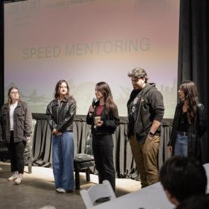 Participants take part in a speed mentoring session, with several speakers standing at the front of the room beneath a “Speed Mentoring” presentation screen.