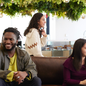 Group of people sitting inside on a couch or standing behind it, talking with each other. A cash register and bakery case behind them.
