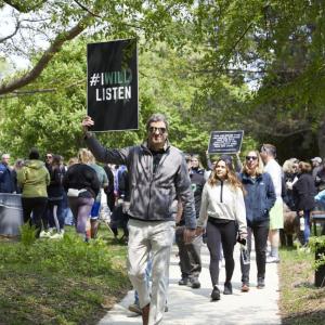 A group walking down a sidewalk and gathered in a park holding a sign "I will listen".