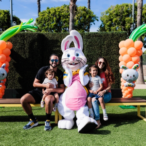 Families sit on a bench posing with a large bunny mascot, surrounded by festive balloon decorations.
