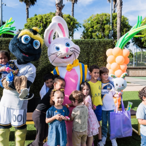 Children gather with two mascots on a turf field, holding gift bags and standing beneath balloon decorations.