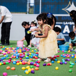 Children collect colorful plastic eggs scattered across an artificial turf field during a community celebration.