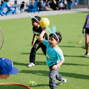 Children participate in a playful outdoor activity on artificial turf, tossing a ball and running between cones and hula hoops.