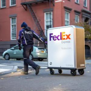 A person in FedEx uniform pulling a tall cart with FedEx Express logo on it crossing a street.
