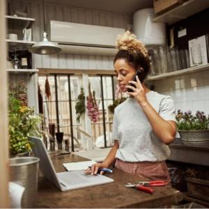 Young woman in a kitchen with her laptop open and speaking on a phone.