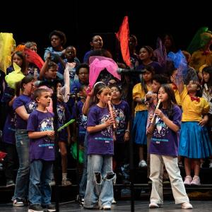 Young musicians perform onstage with recorders while a larger ensemble behind them waves colorful fabric.