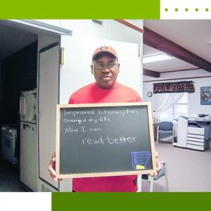 Person stands in a home setting holding a sign that reads "Improving literacy has changed my life. Now I can read better"