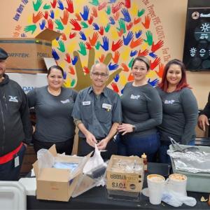 A group of employees pose for the camera, a painted mural of hands on the wall behind them and table of food and serve-ware in front of them.