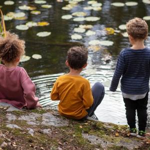 Three small children sitting on the edge of a pond.