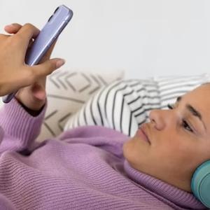 Teenage girl lying down wearing headphones and reading her phone.
