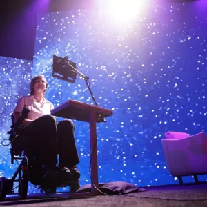 Erin on stage, a digital device in front of them and a colorful starry sky displayed behind.