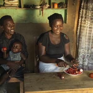 Ernest holds his youngest child in his lap while his wife cuts tomatoes.