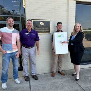 Three men and a woman stand and smile in front of a plaque and around an oversized prop light switch.