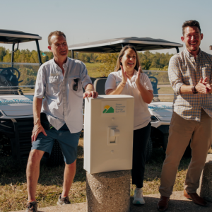 Two men and a woman stand smiling and clapping in front of two UTVs.