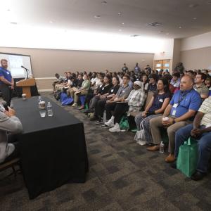 A person speaking at a podium in a room full of seated attendees. A table of other presenters at the front of the room.