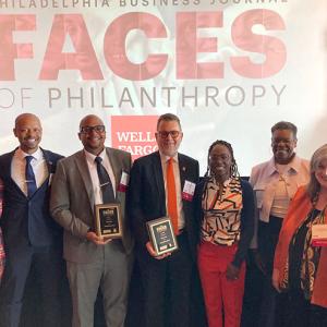 Group standing in front of a 'Faces of Philanthropy' banner. Two people are holding awards.