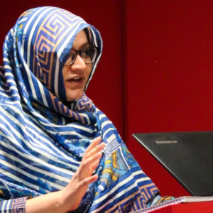 Faiza Yousuf standing at a podium with a laptop. Red walls behind her.