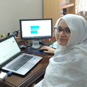 Faiza Yousuf sitting at a home office with a laptop and monitor