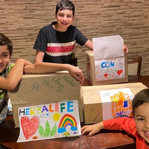 Three children at a table, boxes with Herbalife and drawings on them.