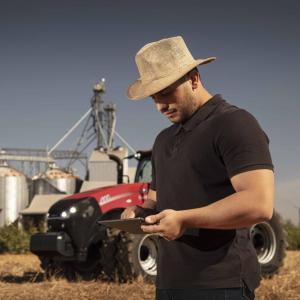 A farmer in a field looking at a tablet device. A red tractor and storage tanks behind them.