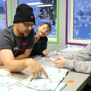 An adult pointing to instructions while three kids assemble a lego toy. Other tables doing the same.