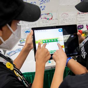 Children pointing at the same tablet, wearing matching lanyards and hats.