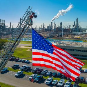 An American flag high above a parking lot with industrial plant behind it.