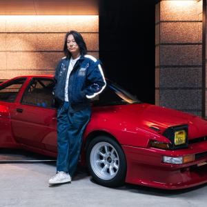 Sung Kang leaning against a red sports car with pop-up headlights in a modern garage, wearing a dark jacket and loose-fit pants.