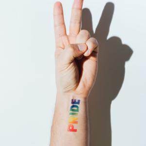 Arm shown with PRIDE written on the wrist in rainbow colors and the fingers in a V for victory sign.