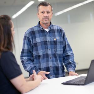 One person sat at a desk with a laptop, talking to a person who is stood beside them