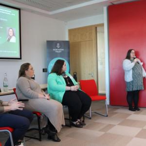 Three seated people at the front of a room. One standing with a microphone speaking to others.