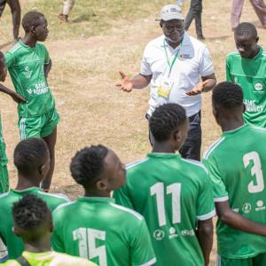 A soccer team huddled and the coach talking with them. They are wearing green uniforms with white writing, standing on a grass and sand field.