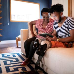 Photo of a woman sitting on a couch next to a man with prosthetic legs, both are looking at a tablet