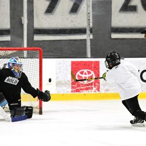 Youth hockey player takes a shot on goal as a goalie in Black, Silver & Bold gear makes a save, with a coach watching on the ice.