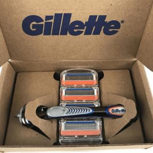 An open cardboard box with Gillette logo on the inside. A razor handle and cartridge refills packaged inside.