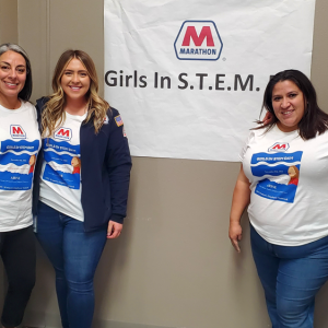 A group of five women with arms around each other in front of a banner "Girls in S.T.E.M." and marathon logo. Each wears a Marathon t-shirt.