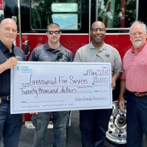 Four people standing in front of a fire truck, holding a large check.