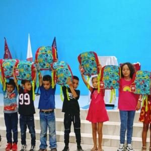 Group of children stood in a line in front of a blue wall holding up matching bags