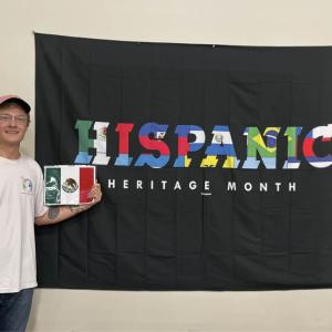a person standing by a "Hispanic Heritage Month" Flag, holding a smaller Mexican flag.