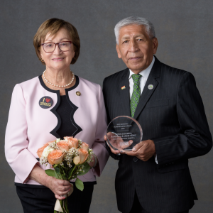 Two people stood next to each other smiling, one person holding flowers and the other holding an award