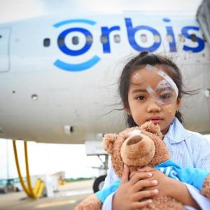Han wearing an eye patch, holding a teddy bear in front of a plane with Orbis on the side.