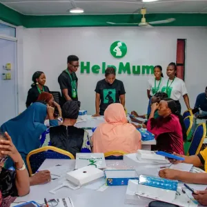 A group of people, some seated, some standing, in a conference room. A speaker at the front. "Help Mum" sign behind them.