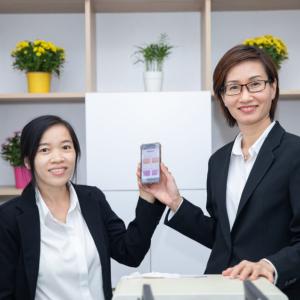 Two people holding a phone up. Shelves behind them each with flowers in them.